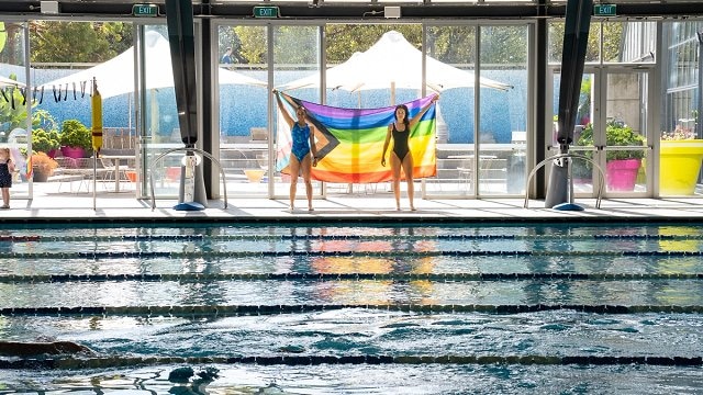 Two females holding the Pride flag next to the indoor pool at Cook + Phillip Pool